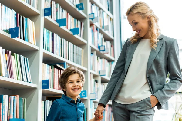 Cheerful woman holding hands with cute kid and standing with hand in pocket — Stock Photo