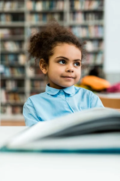 Selektiver Fokus glücklicher afrikanisch-amerikanischer Kinder, die neben Buch auf dem Tisch lächeln — Stockfoto