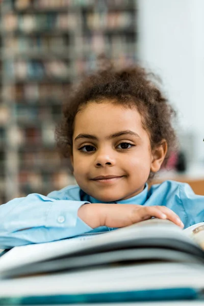 Selective focus of happy african american kid looking at camera near book on table — Stock Photo