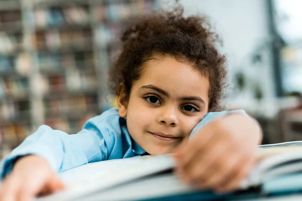 Вибірковий фокус щасливої афроамериканської дитини, яка посміхається, лежачи біля книги на столі — стокове фото
