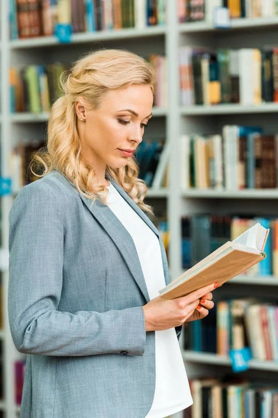 Schöne blonde Frau steht und liest Buch in Bibliothek — Stockfoto