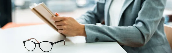 Panorama-Aufnahme einer Brille auf einem Tisch in der Nähe einer Frau mit Buch — Stockfoto