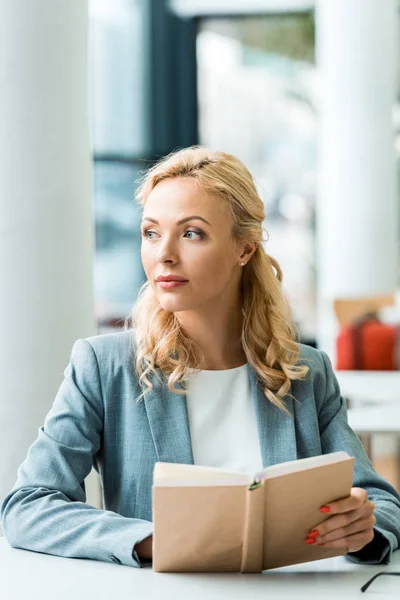 Enfoque selectivo pensativo mujer rubia sosteniendo libro mientras está sentado en la biblioteca - foto de stock