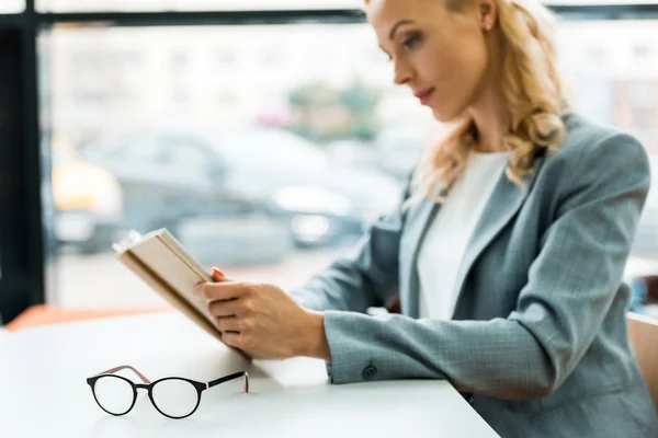 Selective focus of glasses on table near woman reading book — Stock Photo