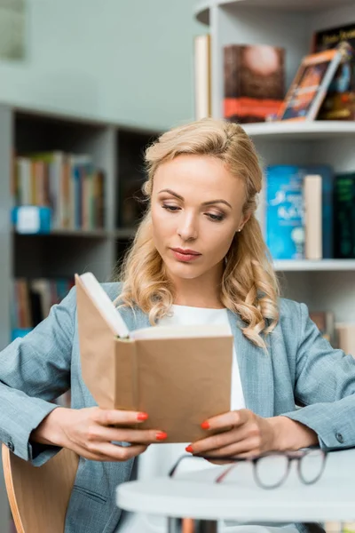 Attraktive und konzentrierte blonde Frau sitzt in der Bibliothek und liest Buch — Stockfoto