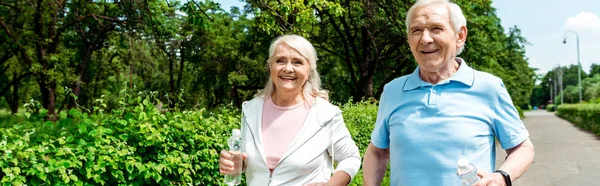 Panoramic shot of happy senior woman holding bottle with water while running near husband in park — Stock Photo