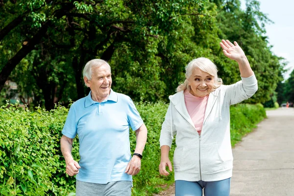 Alegre hombre mayor mirando esposa saludando de la mano en parque - foto de stock