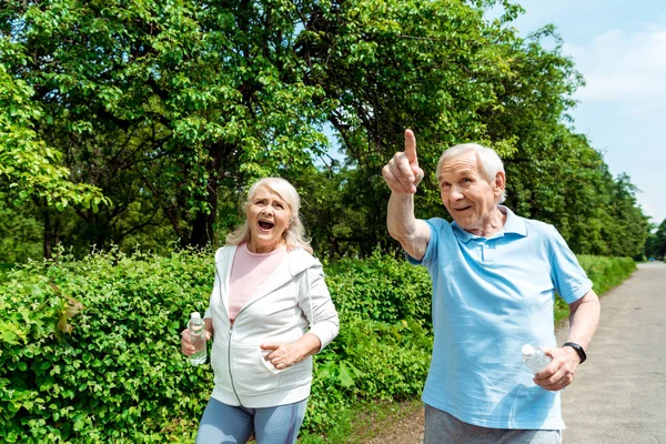 Happy senior man pointing with finger near wife holding bottle in park — Stock Photo