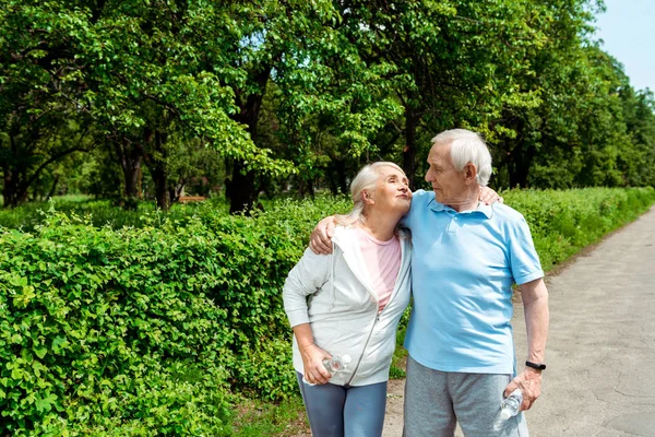 Heureux senior homme regardant femme et tenant bouteille avec de l'eau dans le parc — Photo de stock