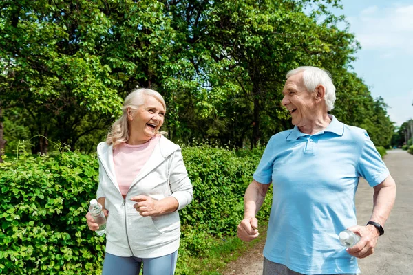 Cheerful woman with grey hair looking at husband while holding bottle and running in park — Stock Photo