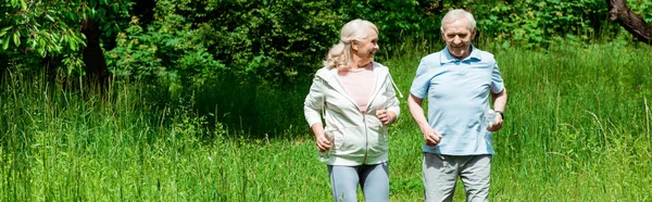 Panoramic shot of senior woman looking at husband while running in green park — Stock Photo