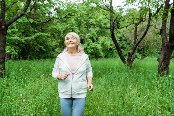 Cheerful senior woman with grey hair running in green park — Stock Photo