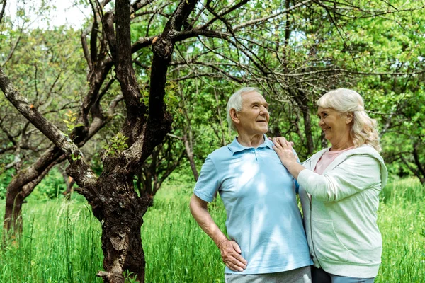 Homme âgé joyeux debout avec la main sur la hanche près de la femme et l'arbre vert — Photo de stock