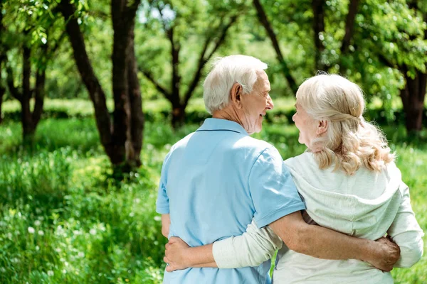 Smiling pensioner hugging happy senior wife while standing in green park — Stock Photo