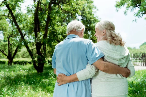 Atrás ver jubilado hombre abrazando senior feliz esposa en verde parque - foto de stock