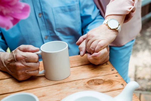 Visão cortada de homem sênior com xícara de chá de mãos dadas com a esposa aposentada — Fotografia de Stock