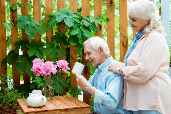 Веселая женщина на пенсии, стоящая рядом с счастливым пожилым мужчиной, глядя на чашку — стоковое фото