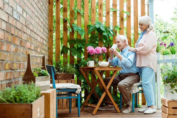 Избирательный фокус веселой старшей жены, стоящей рядом с мужем и пьющей чай — стоковое фото