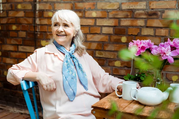 Allegra donna anziana sorridente mentre seduto vicino a tazze e teiera — Foto stock