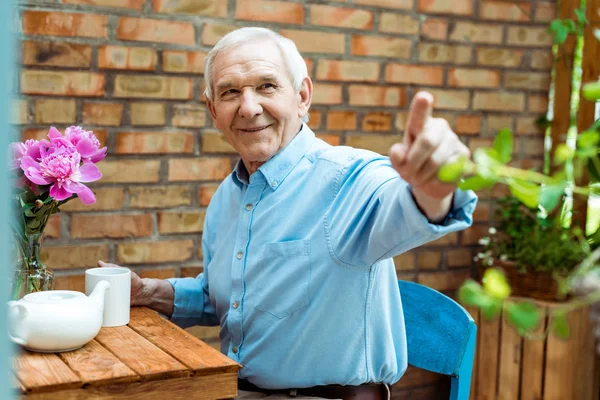 Selektiver Fokus eines glücklichen Seniors, der mit dem Finger zeigt, während er Tasse hält — Stockfoto
