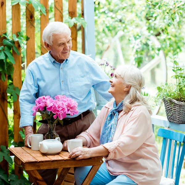 Счастливый старший мужчина стоя и глядя на веселую жену возле чашек — стоковое фото