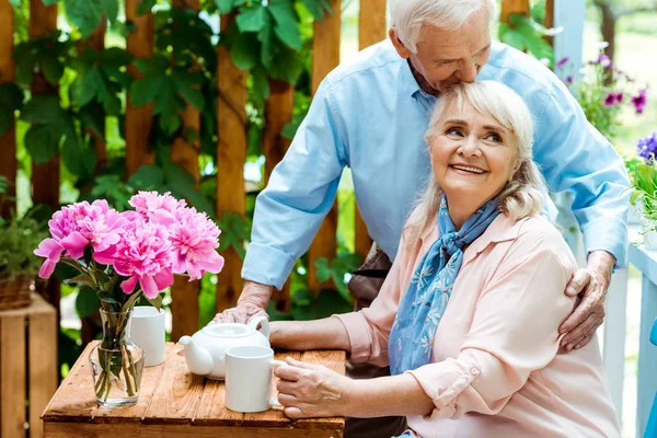 Счастливый пожилой мужчина целует веселую жену сидя рядом с чашками — стоковое фото