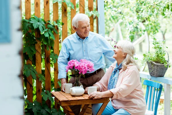 Selective focus of happy senior man looking at retired wife sitting near cups — Stock Photo