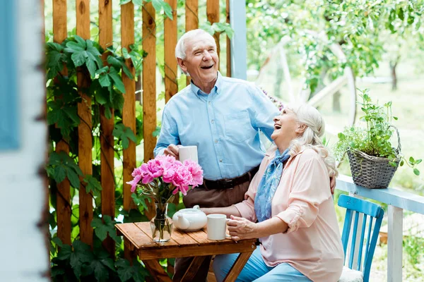 Selective focus of happy senior man laughing with cheerful wife near pink flowers — Stock Photo