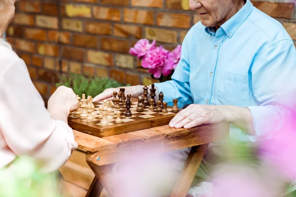 Vista recortada de pareja jubilada jugando ajedrez en mesa de madera - foto de stock