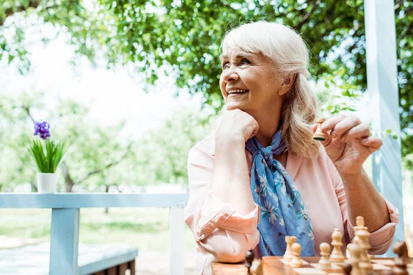 Избирательный фокус веселой пенсионерки, держащей деревянную фигуру во время игры в шахматы — стоковое фото