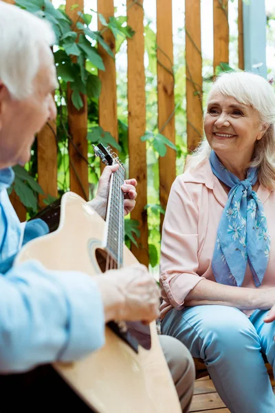 Избирательный фокус счастливой пенсионерки, смотрящей на мужа, играющего на акустической гитаре — стоковое фото