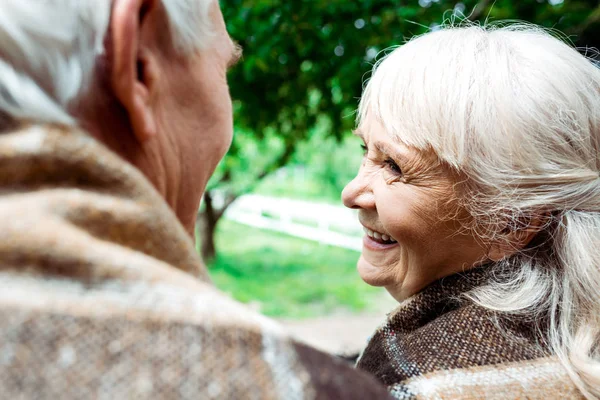 Selektiver Fokus einer glücklichen Seniorin in karierter Decke, die ihren Mann ansieht — Stockfoto