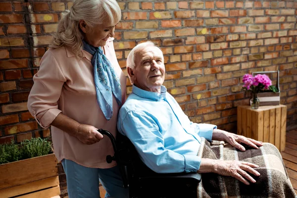 Веселая пожилая женщина, стоящая рядом с мужем-инвалидом в инвалидном кресле — стоковое фото