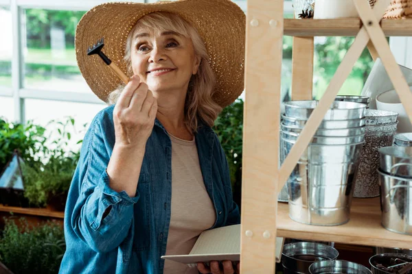 Cheerful senior woman in straw hat holding small rake and notebook — Stock Photo