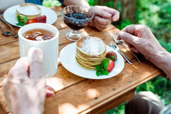 Vista recortada del hombre mayor sosteniendo la taza de té cerca de la esposa y sabrosos panqueques - foto de stock