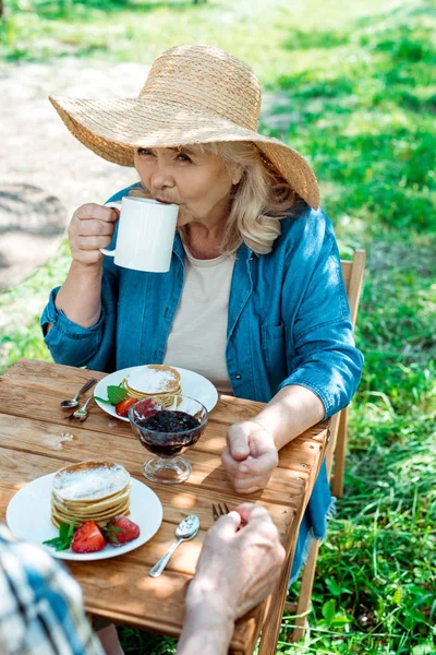 Mulher sênior em chapéu de palha beber chá perto de panquecas doces e saborosas — Fotografia de Stock