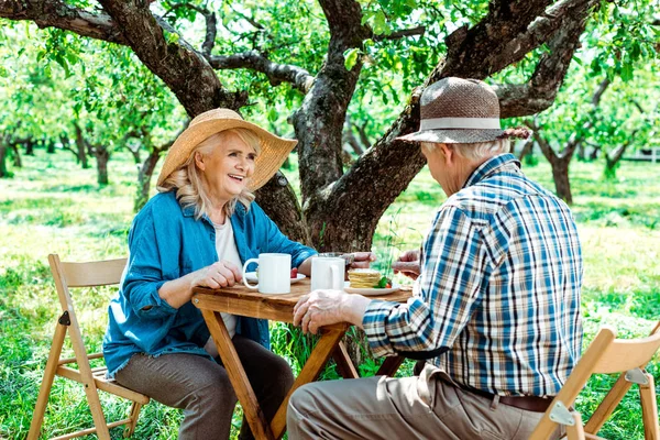 Alegre senior mujer en paja sombrero mirando marido cerca árbol - foto de stock