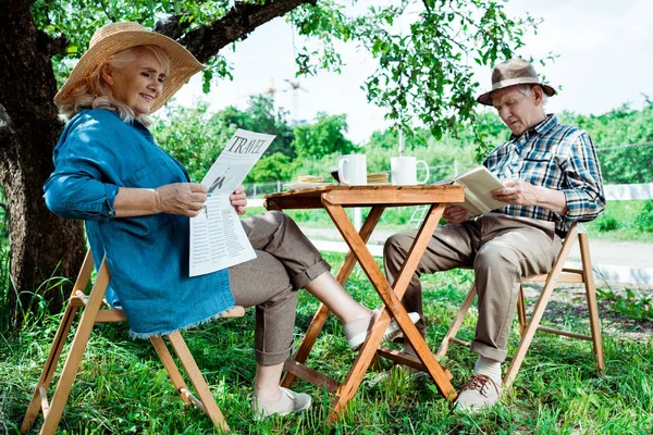Donna anziana che legge giornale di viaggio vicino al marito con libro — Foto stock