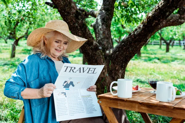 Mujer mayor con sombrero de paja sentada cerca del árbol y leyendo el periódico de viajes - foto de stock