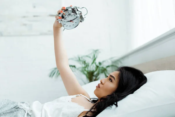 Chateado asiático menina olhando para despertador enquanto deitado no cama — Fotografia de Stock