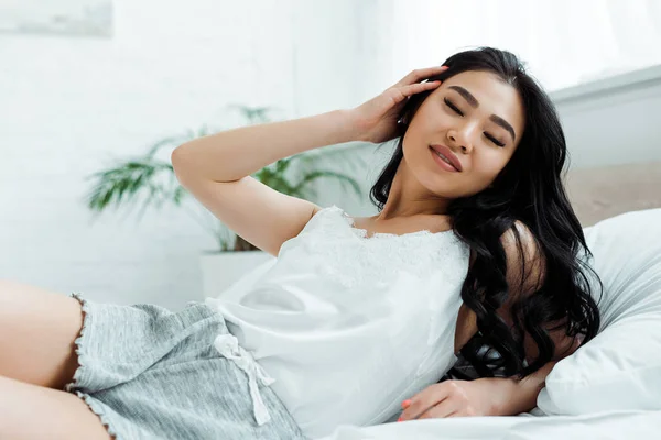 Happy asian girl with closed eyes lying on bed and touching hair — Stock Photo
