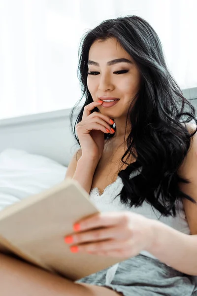 Selective focus of attractive thai woman reading book and touching face — Stock Photo