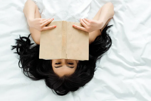 Vue du haut de la fille asiatique couvrant visage avec livre tout en étant couché sur le lit — Photo de stock