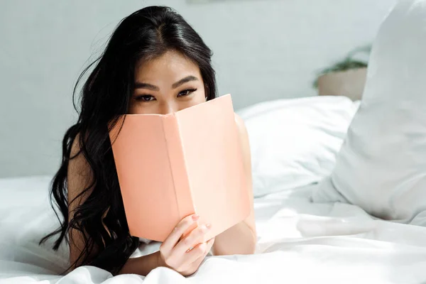 Азіатська молода жінка прикриває обличчя книгою, лежачи на ліжку — стокове фото