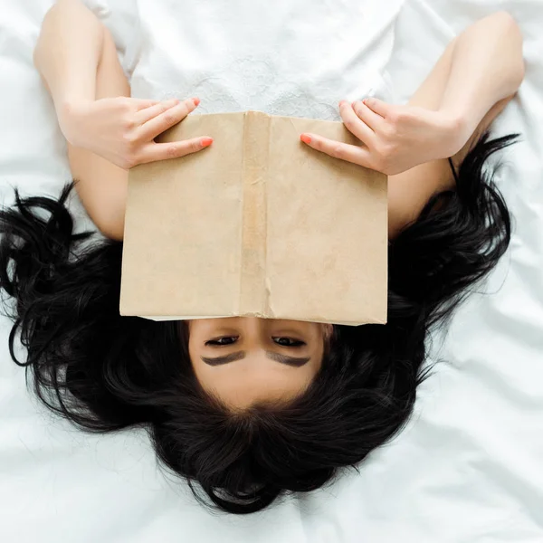 Vue de dessus de la femme thai couvrant le visage avec le livre tout en étant couché sur le lit — Photo de stock