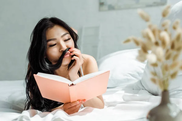 Foco seletivo de menina asiática com rosto de pato tocando o cabelo enquanto lendo livro — Fotografia de Stock