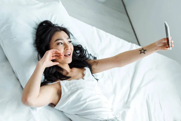 Над головой вид азиатской девушки, делающей селфи на смартфоне, лежащей на кровати — стоковое фото