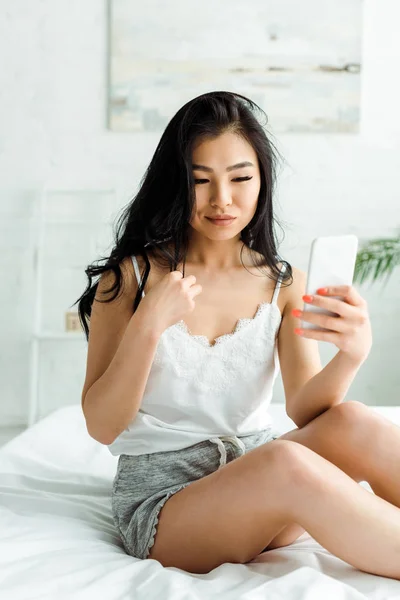Enfoque selectivo de bonita chica asiática usando smartphone en el dormitorio - foto de stock