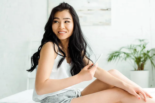Hübsche Thailänderin hält Smartphone in der Hand und lächelt im Schlafzimmer — Stockfoto
