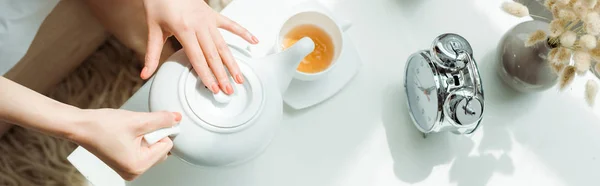 Panoramic shot of woman holding teapot near cup and vintage alarm clock — Stock Photo